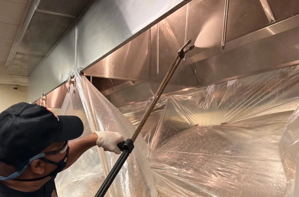 Professional technician cleaning a commercial kitchen grease exhaust hood system with pressure washer and plastic sheeting
